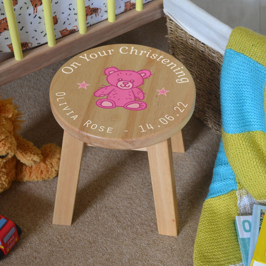 A personalised wooden stool in a children's size. The stool has a design printed on the wooden seat which features white text in a circle around the outside with a pink teddy bear in the middle.