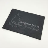 Personalised rectangle slate cheese chopping board pictured on a white background. Engraved with a festive design featuring a Christmas tree and the words, 'The Wilson Family, Christmas Cheese Board'.