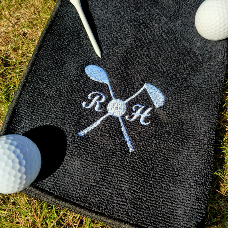 A personalised golf towel in black with white embroidery. The design features two golf clubs and custom initials. 