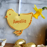 The Easter chick decoration hanging in a kitchen, the Easter decoration is personalised and is made from pastel yellow acrylic and has a white ribbon for hanging up.