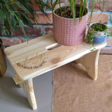 A personalised wooden house plant stand in a rectangle shape. Multiple potted plants are displayed on top and the bench has an engraved name and message. 