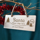 Personalised Oak Christmas hanging sign featuring a festive design. Included are the words 'Santa, please stop here' and it has been personalised with 'for Eva & Daniel' engraved at the bottom. Pictured hanging from a tree by a piece of hessian rope. 