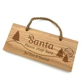 Personalised Oak Christmas Santa sign for children. Featuring a design with Christmas trees and a Santa hat. Included are the words 'Santa, please stop here' along with your added personlisation. Shown here as 'for Eva & Daniel'.
