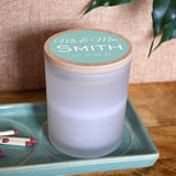 A personalised wedding candle in a glass jar with space for the couple's surname and a date. The design is printed in white lettering on a light blue background.