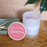 A personalised wedding candle in a glass jar with space for the couple's surname and a date. The design is printed in white lettering on a light pink background.