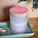 A personalised wedding candle in a glass jar with space for the couple's surname and a date. The design is printed in white lettering on a light pink background.