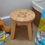 A personalised wooden stool in a children's size. The stool has a design engraved on the wooden seat which features text in a circle around the outside with a teddy bear in the middle.