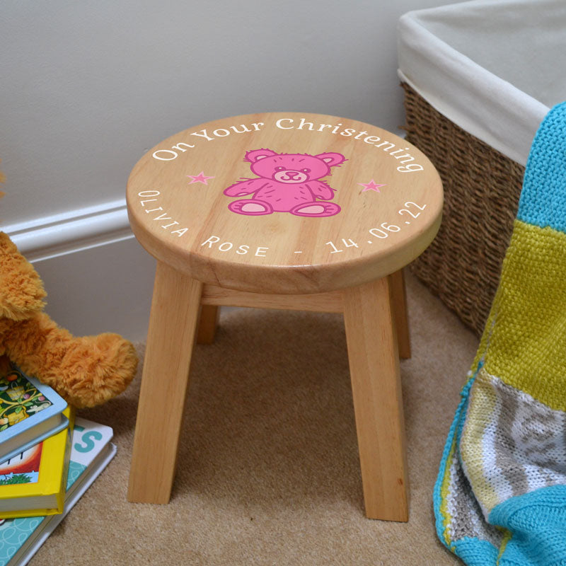 A personalised wooden stool in a children's size. The stool has a design printed on the wooden seat which features white text in a circle around the outside with a pink teddy bear in the middle.