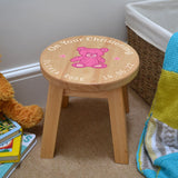 A personalised wooden stool in a children's size. The stool has a design printed on the wooden seat which features white text in a circle around the outside with a pink teddy bear in the middle.