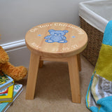 A personalised wooden stool in a children's size. The stool has a design printed on the wooden seat which features white text in a circle around the outside with a blue teddy bear in the middle.