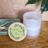 A personalised candle which can have a custom message printed on the bamboo lid. The candle is in a glass jar and the lid features a circle of green leaves with your message printed in green script lettering in the middle.