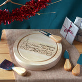 Personalised Christmas cheese board with knife set. Pictured in a festive scene with cheese knifes on display. Engraved on the top of the board is the message "Annie & Martin's Christmas Cheese Board" and cheese illustrations.