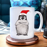 A personalised Christmas mug with a grumpy penguin illustration. The Penguin is wearing a Santa hat with the name "Harry" written on it
