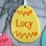 A personalised yellow Easter decoration hanging up in a kitchen. The decoration is egg shaped and has a custom name printed in the centre and a white ribbon.