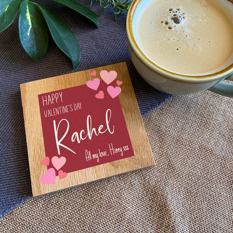 A personalised Valentine's Day coaster. The coaster is square and made from oak with a dark red design printed in the middle on the font. There are light pink hearts and white lettering on the design. 