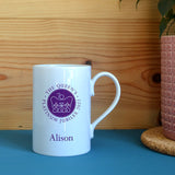 A personalised porcelain jubilee mug in white with the official emblem for the Queen's Platinum Jubilee 2022 printed on the front in purple. 