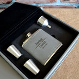 A personalised metal hip flask with a grey leather exterior. The hip flask is in a matching grey leather box with a funnel and two shot glasses.