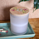 A personalised glass jar candle with a bamboo lid. The bamboo lid has two cherry blossoms and a daisy in the middle with green leaves sprouting from behind. Around the flowers is a message of your choice in pink lettering.