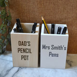 Personalised wooden pencil pot desk tidys pictured containing pens and pencils. One is finished naturally and the other is white. Each engraved with a personal message; "Dad's Pencil Pot" and "Mrs Smiths Pens".