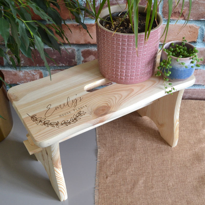 A personalised wooden house plant stand in a rectangle shape. Multiple potted plants are displayed on top and the bench has an engraved name and message. 