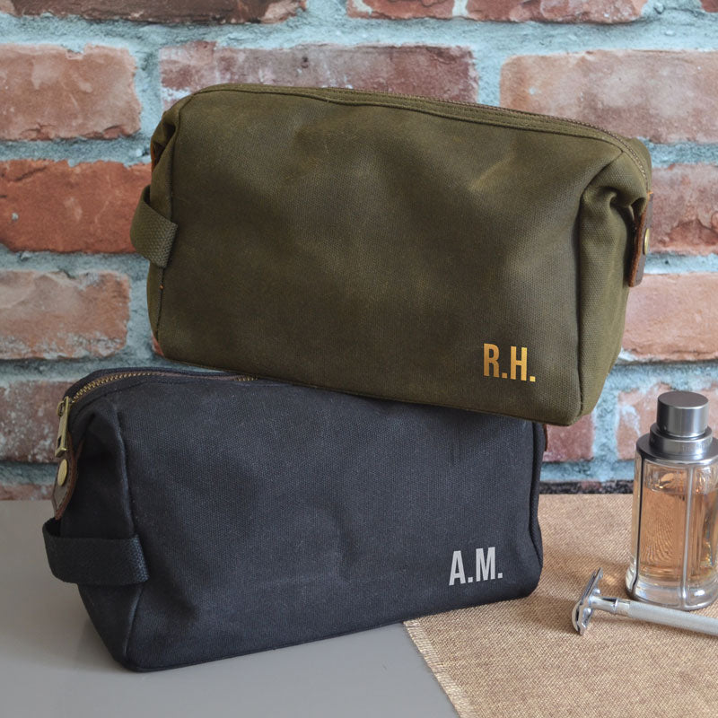 Two personalised waxed cotton wash bags, one in green and one in black. Both wash bags have custom initials printed on the front