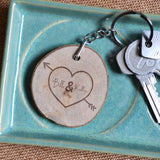 A personalised wooden tree slice keyring with an arrow pierced heart and two names engraved on the front. There is also space to add an optional date. The names and date are enclosed within the heart shape.