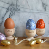 Three personalised wooden egg cups each with a different name engraved on the front, the egg cups hold a mix of chocolate Easter Eggs and boiled dippy eggs. 