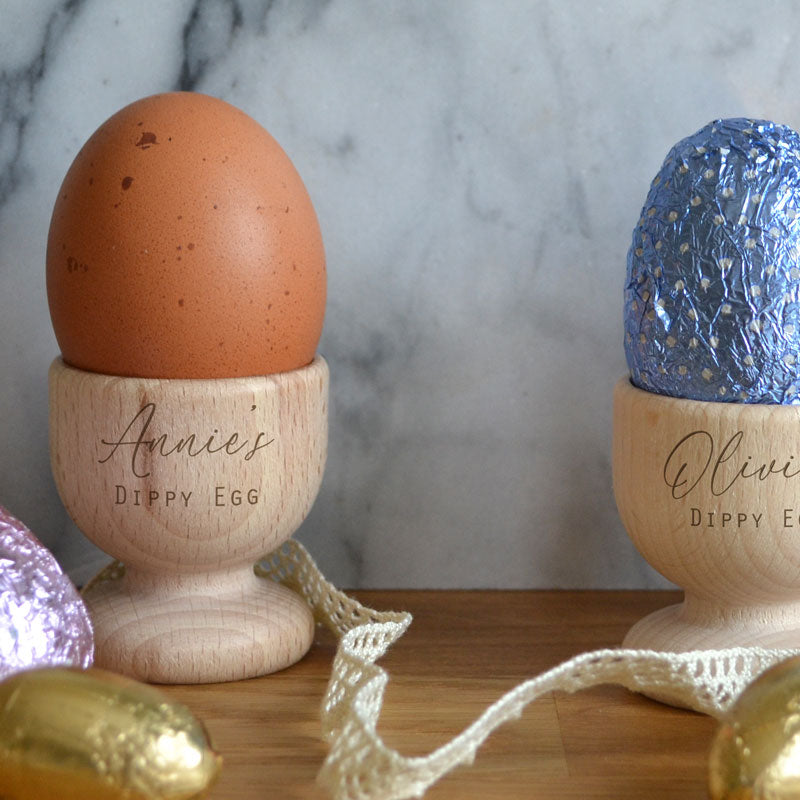 Two personalised wooden egg cups with an engraved design on the front. One egg cup holds a boiled egg and one a chocolate Easter egg. 